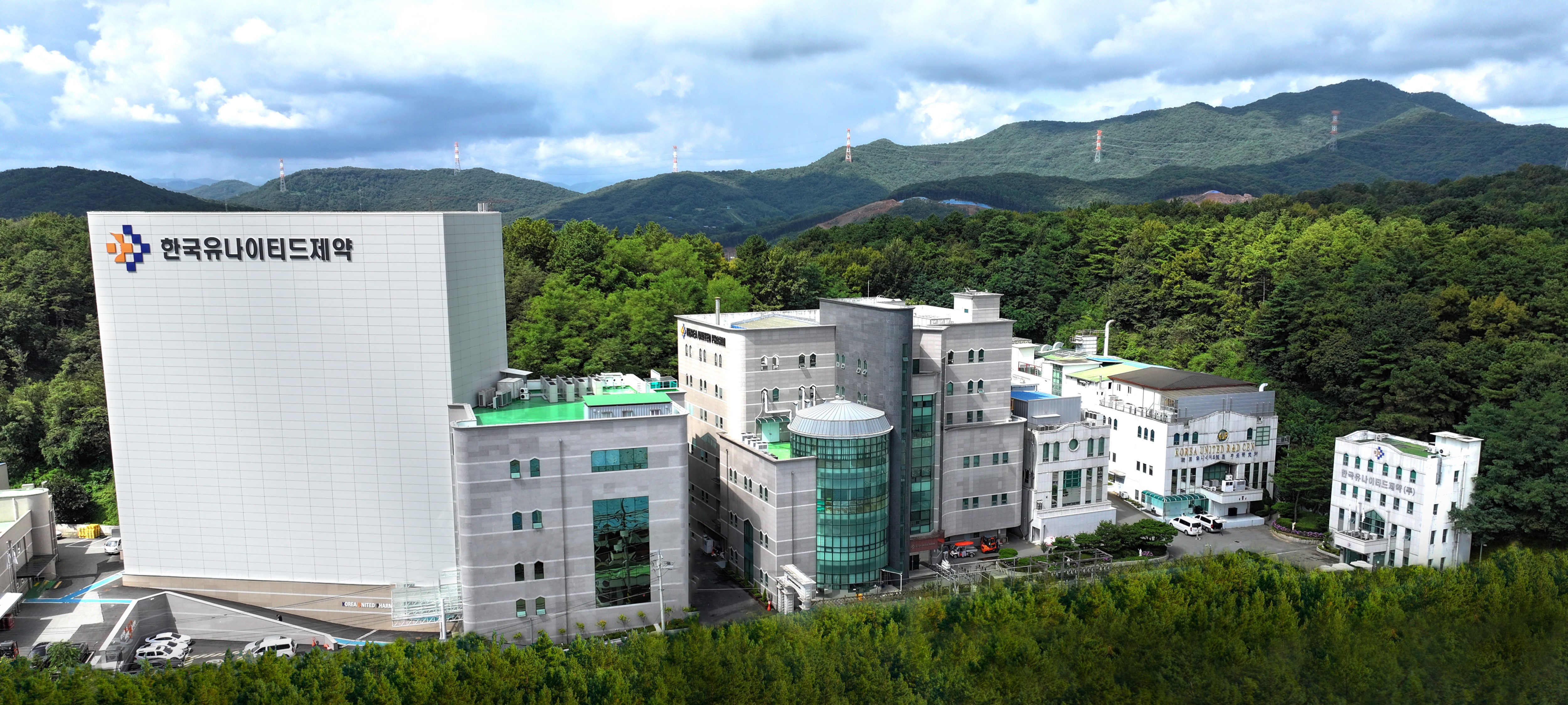 한국유나이티드제약 세종1공장 전경 산과 숲을 배경으로 한국유나이티드제약 로고가 보이는 세종1공장 전경