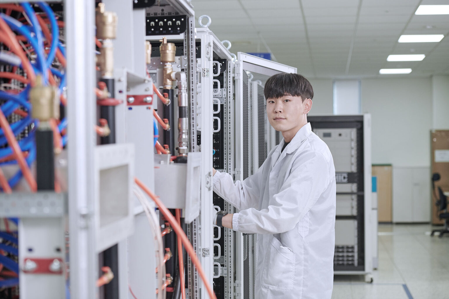 RFHIC 마이크로파 발생기 생산 설비 점검 흰 작업복을 입은 작업자가 케이블이 연결된 대형 장비 랙을 점검하는 생산 시설