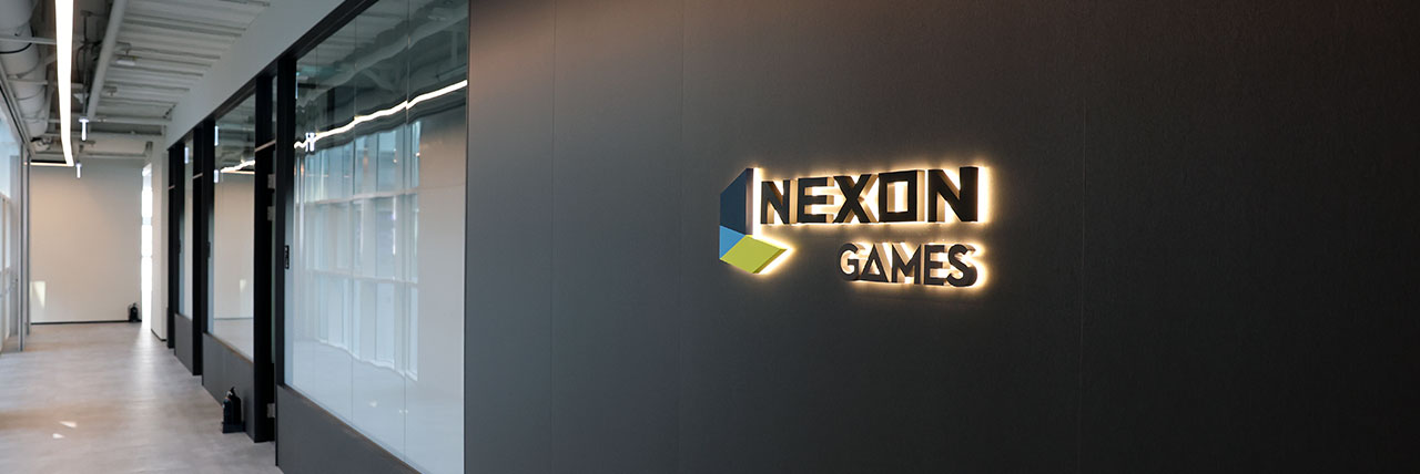 넥슨게임즈 사무공간 입구의 벽면 로고와 복도 검은 벽면의 조명식 NEXON GAMES 로고와 유리 회의실이 보이는 사무실 복도